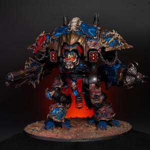 May include: A detailed miniature model of a war machine, featuring a menacing skull-like face with glowing red eyes. The model is predominantly blue and gold, with intricate detailing and a red banner. It has a large cannon and is set on a textured base.