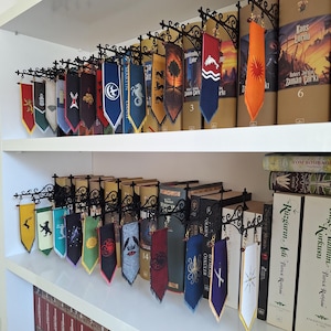 Peut inclure: Une collection de petites bannières en tissu vertical arborant divers symboles et emblèmes graphiques colorés, suspendues à des supports muraux décoratifs en métal noir fixés sur les bords d'étagères blanches remplies de livres reliés.