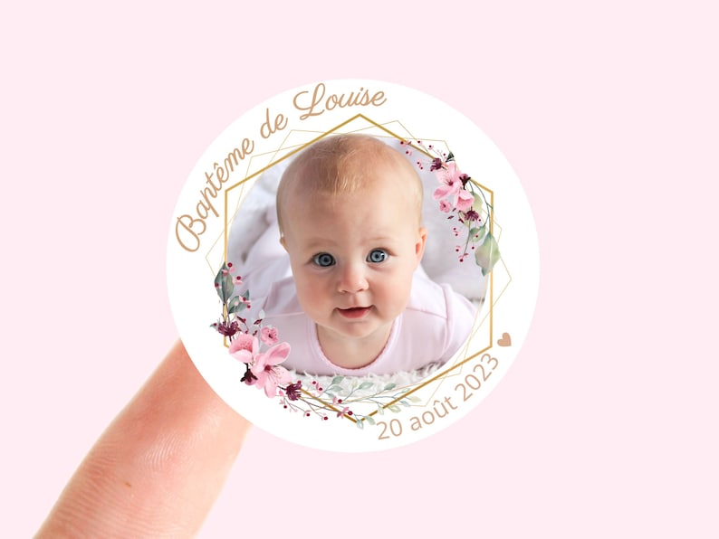 May include: A white round sticker with a gold geometric frame and pink floral accents. The sticker features a photo of a baby girl and the text "Bapt&ecirc;me de Louise 20 ao&ucirc;t 2023".