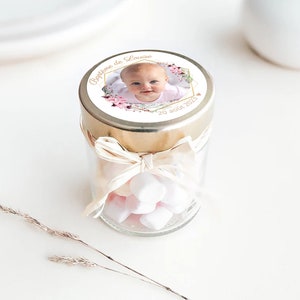 May include: A small glass jar with a gold lid and a white ribbon tied around it. The lid has a round sticker with a photo of a baby girl, pink flowers, and the text "Bapteme de Louise 20 ao&ucirc;t 2023". The jar is filled with white marshmallows.