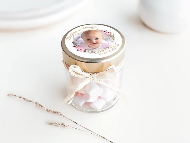 May include: A small glass jar with a gold lid and a white ribbon tied around it. The lid has a round sticker with a photo of a baby girl, pink flowers, and the text "Bapteme de Louise 20 ao&ucirc;t 2023". The jar is filled with white marshmallows.
