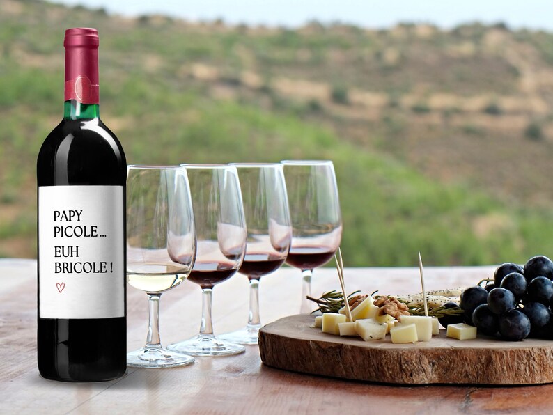 K&ouml;nnte beinhalten: Eine Flasche Rotwein mit einem Etikett, auf dem "Papy Picolo... Euh Bricole!" steht. Die Flasche steht auf einem Holztisch mit vier Weingl&auml;sern, einer K&auml;se- und Traubenplatte und einem verschwommenen Hintergrund aus sanften H&uuml;geln.