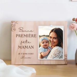Peut inclure: Cadre photo en bois clair avec une photo d'une mère et de son bébé. Le cadre porte l'inscription "Bonne Première Fête Maman" et "Je t'aime fort Louise" en écriture cursive. Motifs floraux gravés aux coins.