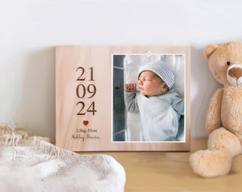 May include: Wooden photo frame with a picture of a newborn baby wearing a blue outfit and a striped hat. The frame has engraved text: date, weight (2.3kg), height (55cm), and a name. A teddy bear is next to the frame.
