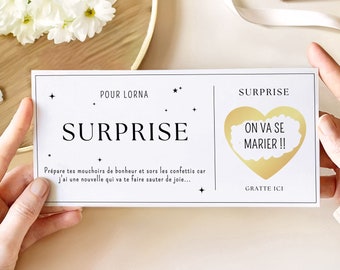 May include: A white card with black text that reads "POUR LORNA" and "SURPRISE". A gold heart with a white cloud inside that reads "ON VA SE MARIER!!" is on the right side of the card. The text "GRATTE ICI" is below the heart.