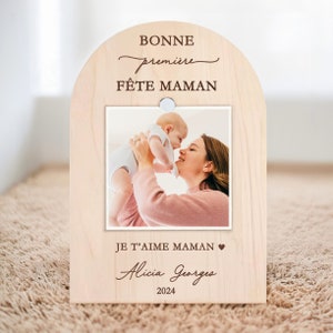 May include: A wooden photo frame with a light brown finish. The frame is shaped like an arch and has a cutout for a photo. The photo shows a mother holding her baby. The frame has the text "BONNE première FÊTE MAMAN" at the top and "JE T'AIME MAMAN" at the bottom. The text "Alicia Georges 2024" is also included.