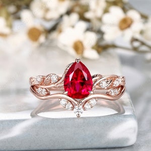 May include: A pear-shaped ruby engagement ring set in rose gold with a delicate leaf design and smaller diamonds. The ring is on a gray surface.