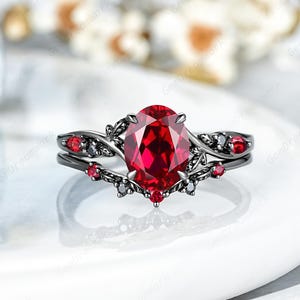 Könnte beinhalten: Ein schwarzes Verlobungsring-Set mit einem großen, oval geschliffenen Rubin-Mittelstein. Der Ring hat kleinere rote und schwarze Akzentsteine und ein dekoratives Blumendesign. Das Set enthält ein passendes Band.