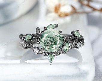 May include: Black ring with a green moss agate gemstone surrounded by smaller green stones and black leaves. The ring is set on a white surface.