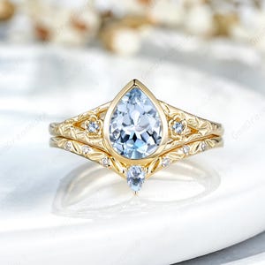 May include: A gold-colored engagement ring set with a pear-shaped, light blue gemstone. The band features floral and leaf patterns, with small accent stones. A matching band sits below the main ring.