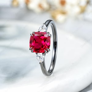 May include: A silver ring featuring a large, cushion-cut, vibrant red gemstone as the centerpiece. Two smaller, pear-shaped white gemstones flank the main stone. The ring's band is a dark, metallic color, creating a striking contrast.