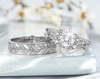 Könnte beinhalten: Zwei silberne Eheringe. Einer hat einen birnenförmigen Diamanten mit floralen Details. Der andere Ring hat ein Band mit einem Blattmuster. Beide Ringe liegen auf einer hellblauen Oberfläche.