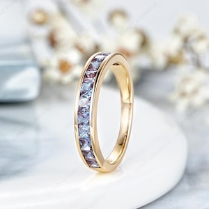 May include: A gold band ring with a row of square-cut, light blue gemstones. The ring is set on a white surface with a blurred background of white flowers. The ring is a piece of jewelry.