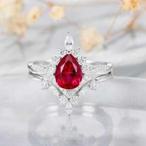 May include: A pear-shaped ruby engagement ring set with a halo of smaller diamonds. The ring is set in white gold and has a delicate, floral design.