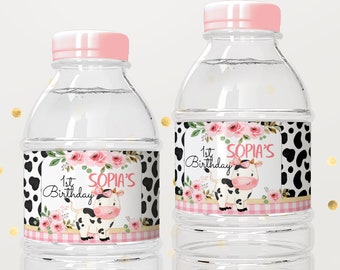May include: Two clear plastic water bottles with pink lids and labels. The labels feature a pink and black cow with flowers and the text "1st Birthday Sophia's".