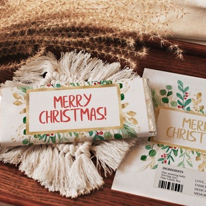 May include: A Christmas-themed candy bar wrapper and card with the text "MERRY CHRISTMAS!" in red and gold. The wrapper and card feature a watercolor design of green leaves, red berries, and gold accents. The candy bar rests on a white macrame coaster.