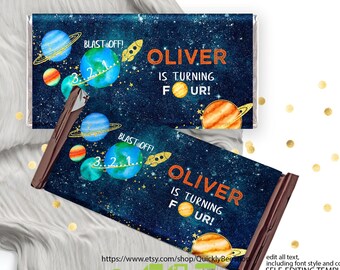May include: Printable chocolate bar wrapper with a space theme. The wrapper features a blue background with stars, planets, and a rocket. The text "Oliver is turning four!" is written in orange and black. The wrapper is designed for a birthday party.