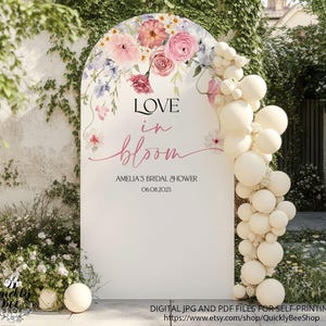 Könnte beinhalten: Ein weißes, bogenförmiges Schild mit Blumenarrangements und Ballondekoration. Auf dem Schild steht "LOVE in bloom" mit dem Text "AMELIA'S BRIDAL SHOWER 06.08.2025". Das Design umfasst rosa, lila und weiße Blumen.