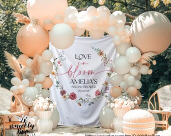 May include: A white backdrop with the text "Love in Bloom Amelia's Bridal Shower 05.06.2026" surrounded by floral illustrations. The backdrop is decorated with a garland of beige balloons.