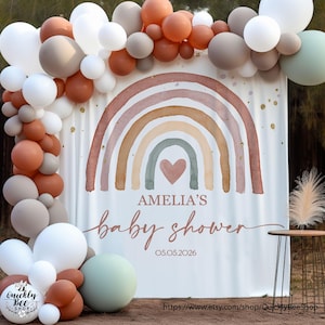 May include: A white backdrop with a watercolor rainbow design in shades of brown, pink, and green. The text "Amelia's Baby Shower" is written in a cursive font with the date "05.05.2026" below. The backdrop is decorated with a garland of balloons in shades of brown, beige, and white.