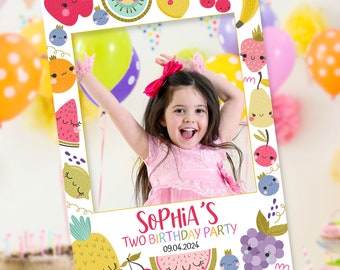 May include: A white photo booth frame decorated with cartoon fruits like cherries, lemons, and watermelon. The frame has the text "Sophia's Two Birthday Party 09.04.2024" and a website address. A young girl is inside the frame, smiling.