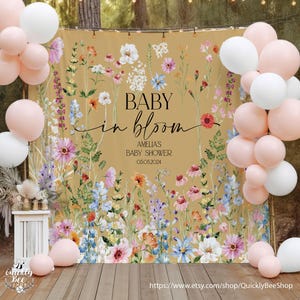 May include: A floral baby shower backdrop with the text "BABY in bloom". The backdrop features a variety of colorful flowers and foliage. White and pink balloons are arranged on either side of the backdrop, creating a festive atmosphere.