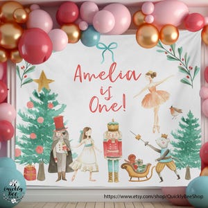 Puede incluir: Un telón de fondo festivo para una celebración de primer cumpleaños, con el texto "Amelia is One!" e ilustraciones de un árbol de Navidad, una bailarina y personajes de Cascanueces. El telón de fondo está rodeado de globos en tonos rosa, rojo y dorado.
