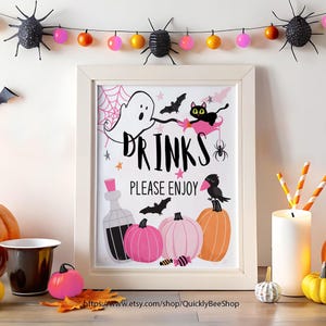 Könnte beinhalten: Ein gerahmtes Halloween-Schild mit dem Wort "DRINKS" und dem Satz "PLEASE ENJOY". Das Schild zeigt Illustrationen eines Geistes, einer schwarzen Katze, Fledermäusen, Kürbissen und einer Spinne. Eine Lichterkette und dekorative Kürbisse befinden sich im Hintergrund.