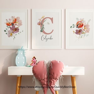 May include: Three white framed prints with watercolor floral designs and a ladybug. The middle print features the letter "C" in a pink color with the name "Calynda" written below it.