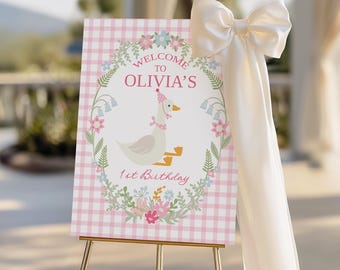 Puede incluir: Un letrero de bienvenida a cuadros rosa y blanco para el primer cumpleaños de Olivia, con un ganso de dibujos animados con un gorro de fiesta y una corona floral. El letrero está sobre un caballete dorado, adornado con un gran lazo crema.
