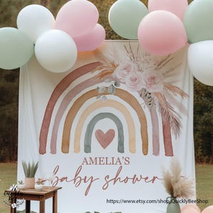 Könnte beinhalten: Ein weißer Hintergrund mit einem Aquarell-Regenbogen in Braun-, Rosa- und Grüntönen. Der Regenbogen hat ein Herz in der Mitte. Der Text "Amelia's Baby Shower" ist in brauner Schrift unter dem Regenbogen geschrieben. Der Hintergrund ist mit weißem Pampasgras und Grünpflanzen dekoriert.