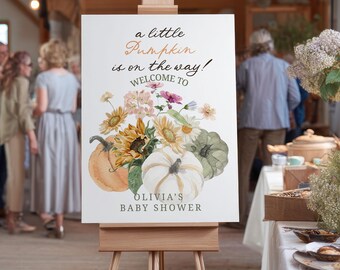 May include: A white sign on a wooden easel with the text "a little Pumpkin is on the way! Welcome to Olivia's Baby Shower." The sign features watercolor illustrations of pumpkins, sunflowers, and other flowers.