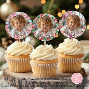 May include: Three cupcakes with white frosting are topped with circular toppers featuring a child's photo and the text "Olivia is One." The toppers are decorated with festive elements. The cupcakes sit on a wooden base.