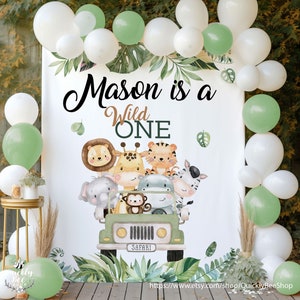 May include: A white backdrop with green leaves and a green jeep with animal characters. The text "Mason is a Wild One" is written in black on the backdrop.  The backdrop is decorated with green and white balloons.