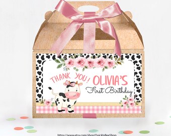 May include: A brown favor box with a pink ribbon and a pink and white cow design. The box has a label that says "Thank You! Olivia's First Birthday".