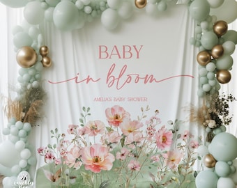 May include: A white backdrop with the text "BABY in bloom" and "Amelia's Baby Shower" in pink script. The backdrop is decorated with a green and pink floral design and a garland of sage green balloons with gold accents.