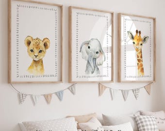May include: Three framed watercolor prints featuring a lion cub, an elephant, and a giraffe. Each animal is centered on a white background with a dotted border, set in a light wood frame. A decorative banner hangs below.