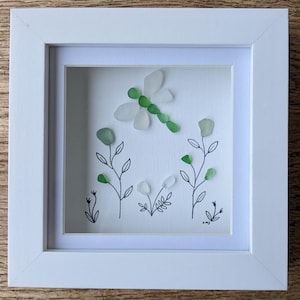 May include: A framed artwork with a dragonfly and floral design. The dragonfly is made from green and clear sea glass, with a green body and clear wings. The artwork includes hand-drawn stems and leaves, with green and clear sea glass accents. The frame is white.