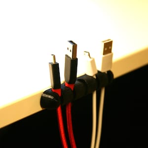 May include: A black cable organizer with four cables running through it. The cables are red, black, white, and white. The organizer is mounted on a white surface.