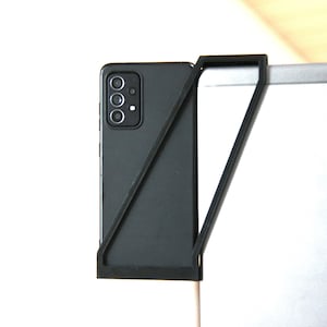 Könnte beinhalten: Ein schwarzes Smartphone mit einem dreifachen Kamerasensor wird in einem schwarzen Plastikständer gehalten. Der Ständer ist an einem silbernen Laptop befestigt.