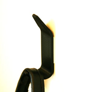 Könnte beinhalten: Ein schwarzer Plastikhaken, der an einer weißen Wand befestigt ist. Ein schwarzer Lederriemen hängt an dem Haken.