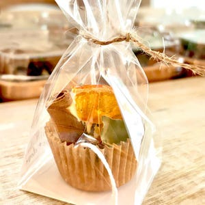 May include: A small gift bag with a clear cellophane wrapper tied with twine. Inside is a brown paper cupcake liner filled with dried orange slices, cinnamon sticks, and green leaves. The background is blurred.