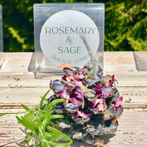 May include: Rosemary & Sage Smudge Cone with a white wax wick. The cone is made of dried herbs and flowers, including rose petals, and is packaged in a clear plastic box.