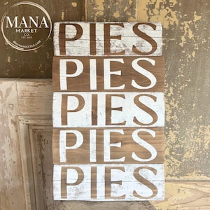 May include: A rustic wooden sign with the word "PIES" repeated five times in white paint. The sign is distressed and has a weathered look.