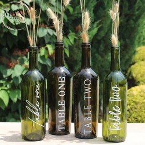 May include: Four green glass wine bottles with white text labels. The labels read "Table One", "Table Two", and "Table one" twice. The bottles are filled with dried grasses.