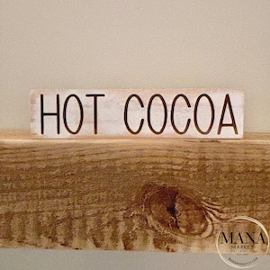 May include: White wooden sign with black lettering that reads "HOT COCOA".