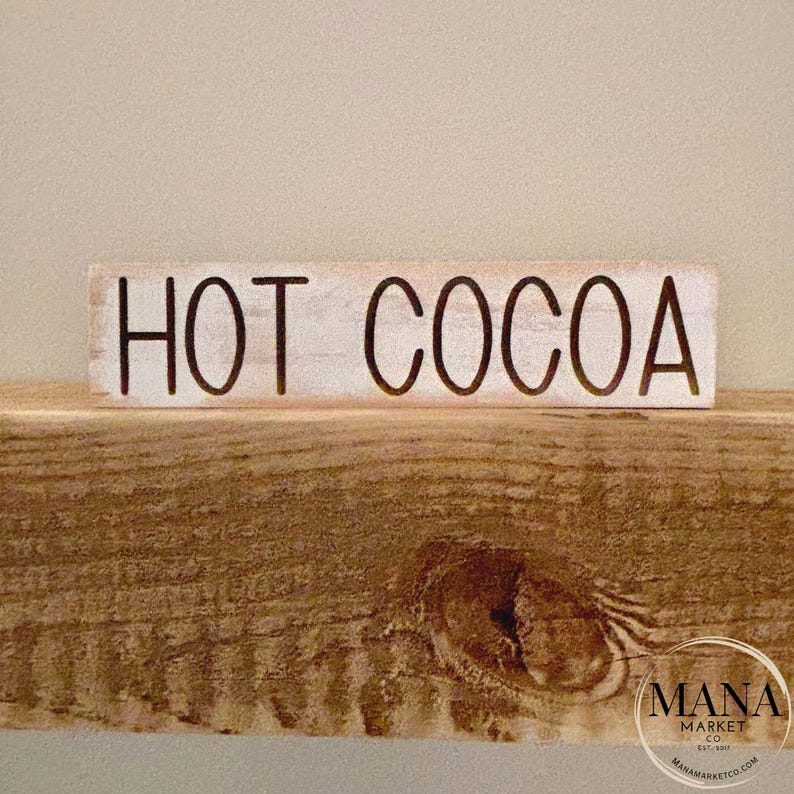 May include: White wooden sign with black lettering that reads "HOT COCOA".