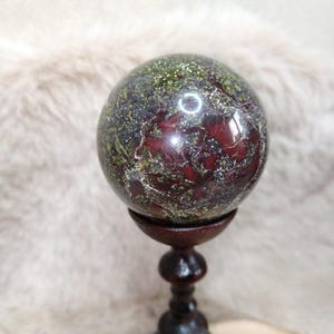 Op de afbeelding: Een gepolijste stenen bol met een gemarmerd patroon van diep bordeauxrood, olijfgroen en metallic zilveren spikkels. De bol rust stevig op een donkerbruine, gedraaide houten voet met een uitlopende basis en een afgeronde bovenkant.