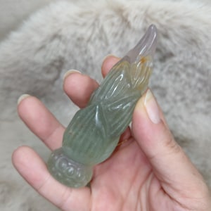 Könnte beinhalten: Eine geschnitzte Figur aus grünem Stein in Form einer Zikade mit einem durchscheinenden, spitz zulaufenden Ende und einer abgerundeten Basis mit kreisförmigen Mustern. Der Stein zeigt natürliche Farbvariationen in Hellgrün, Grau und Bernsteintönen.