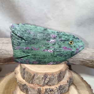Può includere: Una scultura di pesce in pietra scolpita nei toni del verde e del rosa, con un occhio color oro. Il pesce è esposto su una pila di fette di legno, mostrando i suoi colori e le sue trame naturali. La scultura è un oggetto decorativo unico.